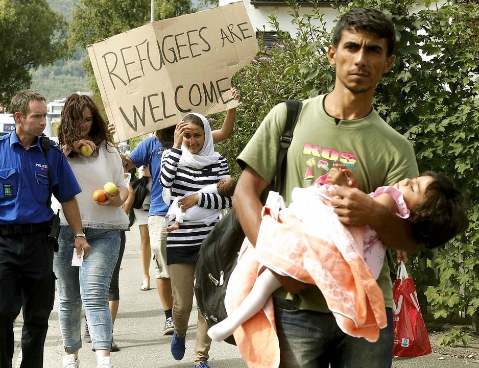 An image of people walking in a protest, carrying babies and carrying a sign that says "Refugees are Welcome". They are accompanied by a police officer.