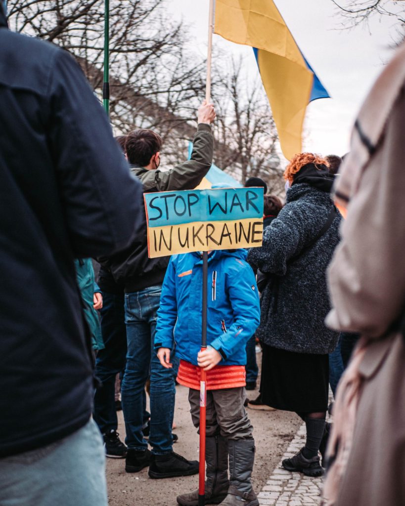 Child dressed for cold weather with sign in front of face reading Stop War In Ukraine surrounded by adults.