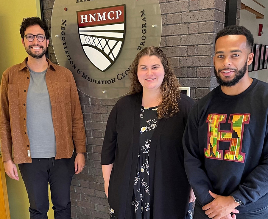 Two men and a woman in their 20s stand in front of the HNMCP logo
