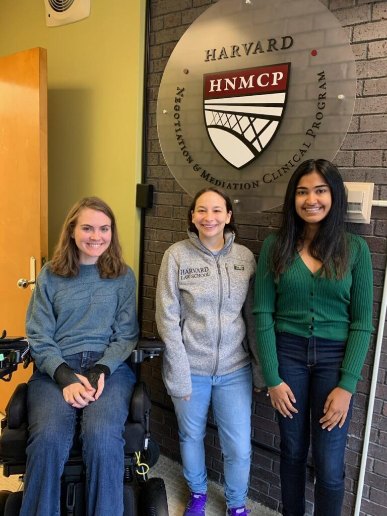 Three women, smiling, in front of the HNMCP logo. Two are standing, one is in a wheelchair.