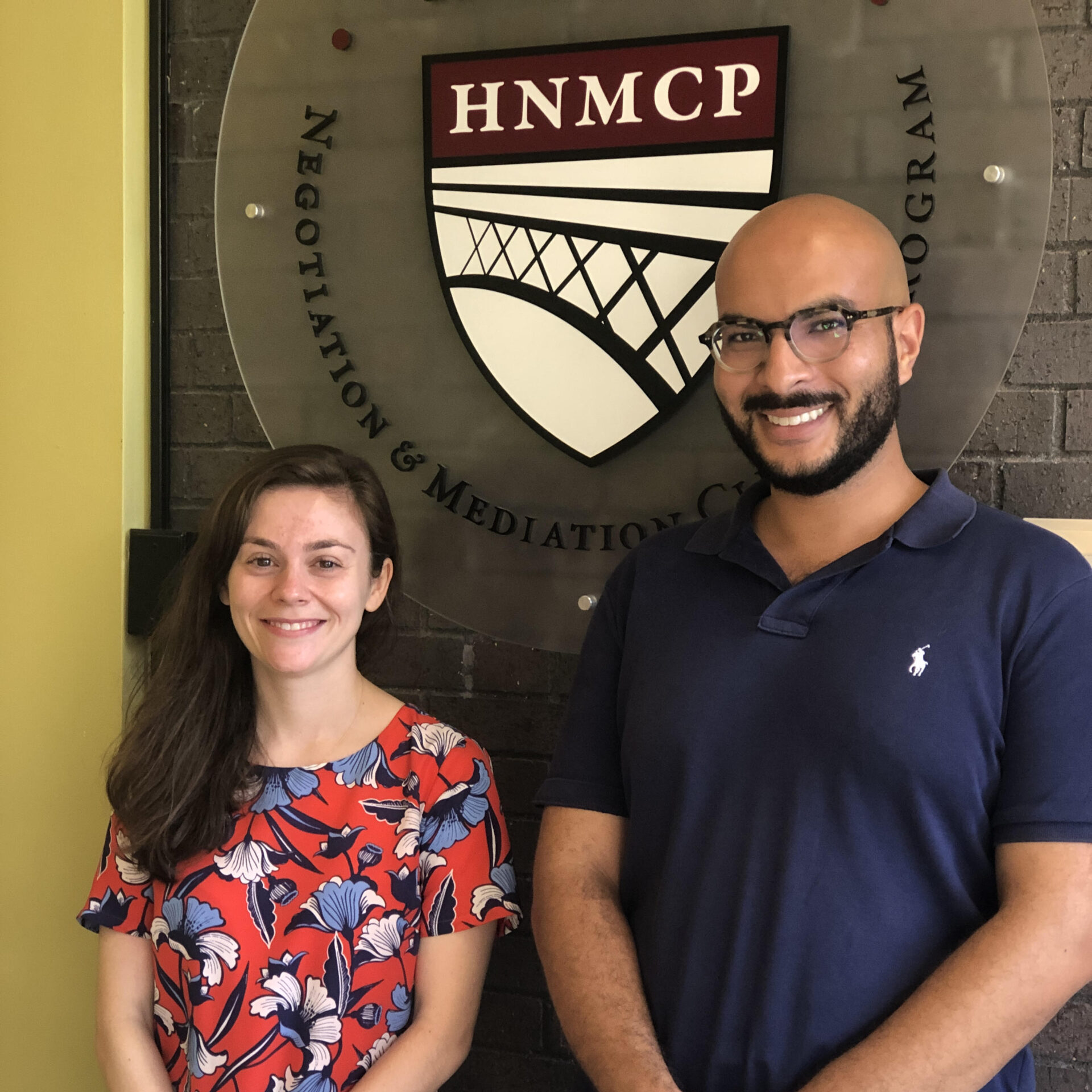 A shorter woman and a taller man, both smiling, standing in front of the HNMCP Logo