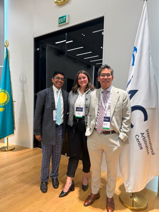 Three colleague, two men and a woman, smiling in front of a flag with International Arbitration Center on it.